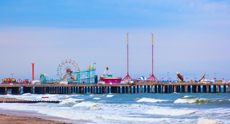 A colorful side view image of the pier in Point Pleasant Beach NJ protruding out over the water with about 10 rides on the pier including a ferris wheel with multi colored seats. Clicking on this image will bring you to the ultimate NJ beach guide