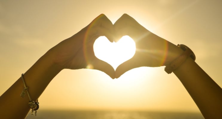 Image of a person making a heart shape with their hands with the backdrop of a twilight sun in the sky over the ocean