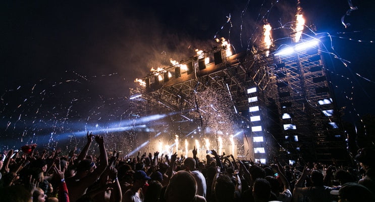 A very fun and exciting up-close photo of an indoor venue showing a band on the stage and fans with their arms up at an exciting concert in NJ