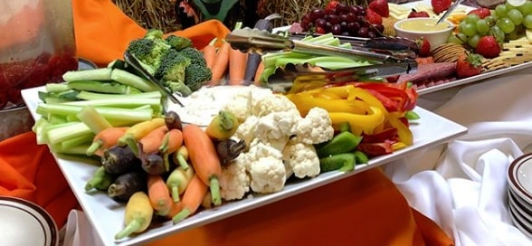 Image of a platter of vegetables on a table with an orange tablecloth including fresh sticks of celery, broccoli florets, carrots, sliced green bell peppers, green grapes and cauliflower with two different dips in the middle