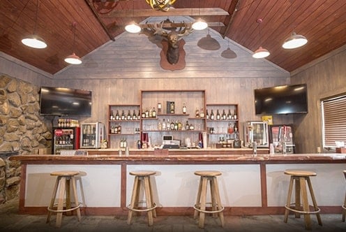 Image of the Pine Ridge Dude Ranch silver dollar bar in light tan and white colors with 4 stools and bottles of alcohol behind the bar