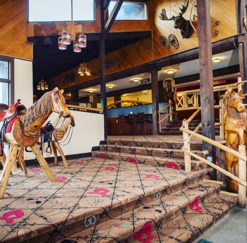 Image of the Pine Ridge Dude Ranch vacation destination just entering the ranch inside with two sets of steps and three wooden horses and a moose head on the pinewood walls in the background